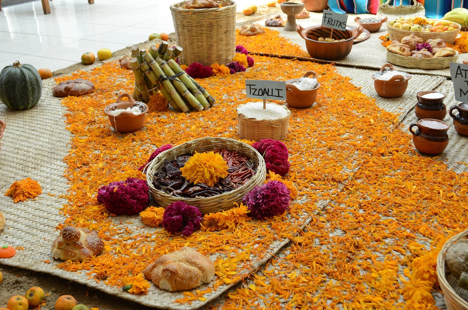 An ofrenda with orange cempasúchil petals spread across woven mats around bread, sugar cane, clay pots and flowers